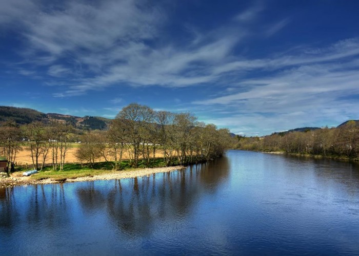 Fishing Spot close to Pitlochry and Fonab Caravanpark. Showing a large river bend with deep blue water and blue skies. A river band of trees and light gravel on the otherside of the river.