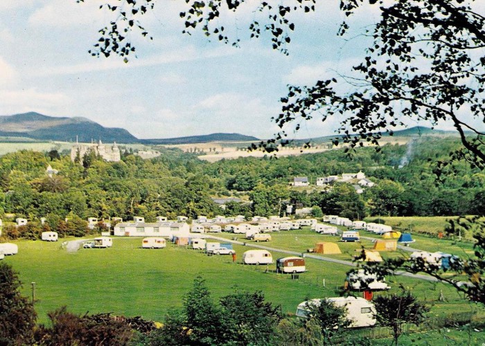 Colourful view of Fonab from throught the trees. Rows of Caravans dating back to the 70's, colourful yellow and blue tents are pitched on the grass area. Rolling hills with greenery in the background